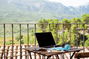 A laptop, blue headsets, a notebook, and a cellphone on the table on a balcony with a beautiful view